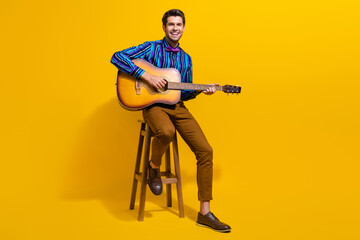 Full body photo of cheerful pleasant man wear stylish shirt sit on stool play on acoustic guitar isolated on yellow color background - obrazy, fototapety, plakaty