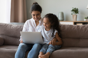 Cute little girl using laptop sit on sofa with loving mom, parent teach child to use modern device...