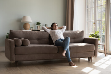 Inside clean cozy light living room young Indian woman resting on sofa put hands behind head closed eyes enjoy day off and fresh conditioned air at modern house. Relaxation, weekend, leisure concept - obrazy, fototapety, plakaty