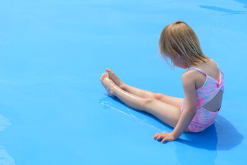 One little blond girl is sitting in the blue pool. A sadness of one little girl in the middle of the summer