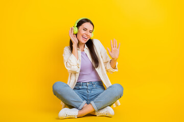 Full size portrait of pretty cheerful girl sit floor hand touch headphones dancing isolated on yellow color background