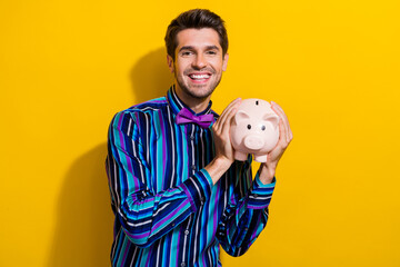 Portrait of toothy beaming nice man with bristle wear vintage bow tie hold piggy money box in arms isolated on yellow color background