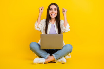 Full size photo of delighted pretty girl sit floor use netbook raise fists triumph luck isolated on yellow color background