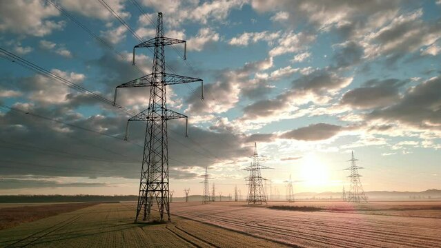 Electricity transmission towers in field with dramatic cloudscape at sunrise aerial shot. Sunlit high voltage poles and energy power lines with nature scenery