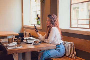 Smiling woman looking at her phone while she is working from a restaurant with a laptop