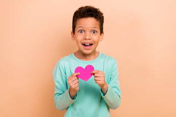 Photo of charming cheerful boy wear trendy turquoise clothes hands hold heart shape paper card isolated on beige color background