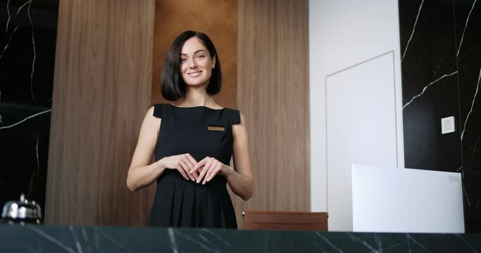 Portrait of elegant Caucasian professional manager of hotel at reception of fancy hotel looking at camera and smiling. Friendly and polite woman working as receptionist wearing black dress.