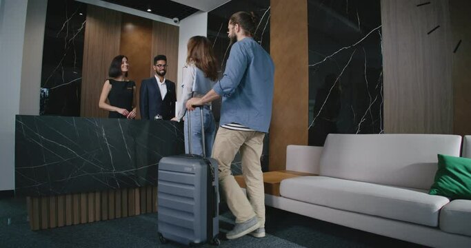 Young lovely couple of man and woman approaching hotel reception with male and female receptionists welcoming new guests. Two attractive people with luggage checking in.