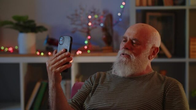 An elderly man in good health and mood uses a smartphone, has video call conversation. A human waves hand as greeting sign and congratulates his interlocutor on the Christmas or New Year holiday