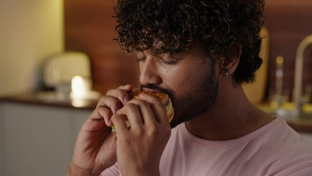 Concept of unhealthy food delivery. Hindu guy enjoying his hamburger. Young man with kinky hair biting tasty burger in kitchen. Male student eating big sandwich