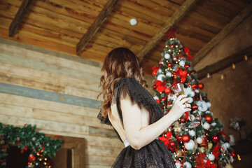 beautiful slender young brunette woman in black evening dress, stands next to decorated Christmas tree. Young brunette woman in a fashionable outfit in the interior. new year party