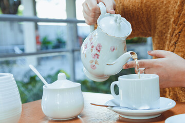 close-up of unrecognizable woman with porcelain teapot pouring tea into porcelain cup on the table - obrazy, fototapety, plakaty