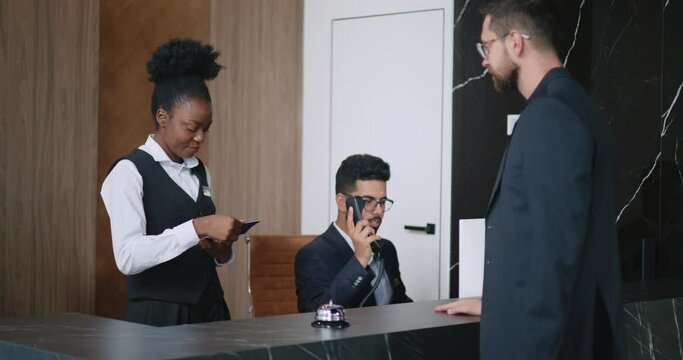 Successful attractive man checking into five-star hotel and giving his passport to friendly African woman working as receptionist. Polite busy manager speaking on phone in the background.