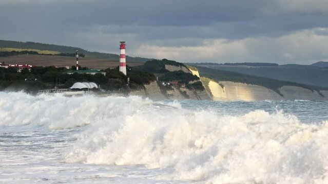 A large wave, when approaching the shore, twists beautifully and the crest of the wave in the foam falls down. In the background there is a steep bank and a high lighthouse.