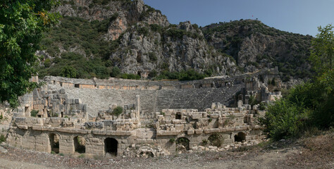 Naklejka premium Glued wide panorama of amphitheatre ancient ruins: carved windows, passages, arches. Remains of ancient Lycia civilization, Myra dead antique city in Turkey. Mountain landscape in the background