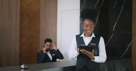 Portrait of beautiful young professional receptionist using tablet online looking at camera and smiling. Busy luxury hotel manager talking on phone taking calls on background.