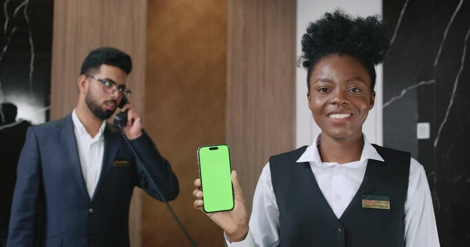 Portrait of African-American hospitable happy female receptionist holding smartphone with green screen chroma key. Handsome Indian male manager talking on phone. Luxury hotel concept.