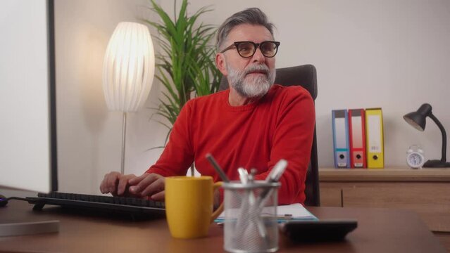 Successful mature guy project manager sitting at workdesk at office, typing on pc computer keyboard and smiling, chatting with clients, enjoying his job, copy space. Freelancer working from home