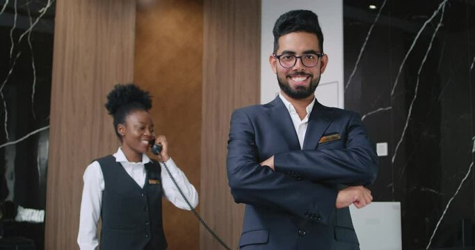 Portrait of handsome happy luxury hotel receptionist in suit looking at camera and smiling. Professional African-American hotel staff member taking calls from customers. Customer care concept.