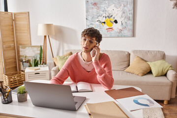 curly androgynous freelancer in pink sweater and eyeglasses talking on smartphone near laptop