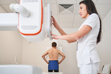 Adult man standing topless next to x-ray machine while female nurse adjusts it