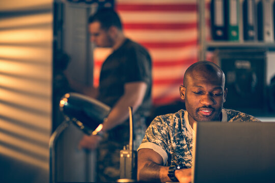 American soldier using a laptop while deployed in an army base with his roomate behind him
