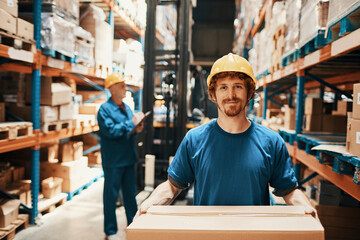 Portrait young man in helmet holding box standing in warehouse looking at camera