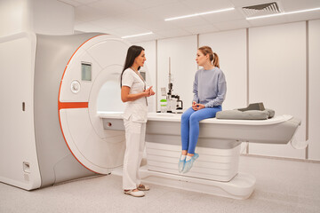 Radiologic technician smiling and talking to female patient sitting on CT scan bed