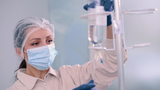 Nurse in protective mask and surgical cap preparing IV bag for intravenous infusion in hospital. Close up of face.