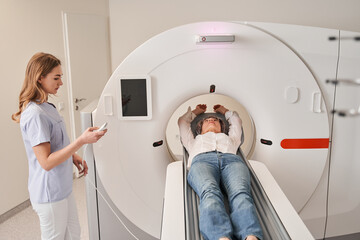 Senior lady laying on CT scan bed while female radiologic technician turning on panel control