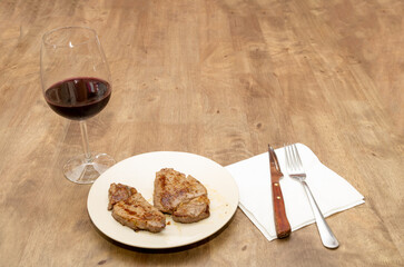 a couple of pieces of grilled beef tenderloin, entrecote on a white plate with a white napkin, a knife and a fork on a wooden table and a glass cup of red wine
