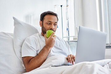 young indian man having video call and holding green apple while lying in hospital bed, healthcare
