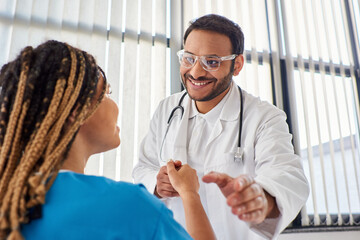focus on jolly indian doctor in glasses shaking hand with his blurred african american patient