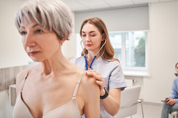 Calm aged woman standing and professional female general practitioner using stethoscope