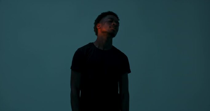 Plain dark studio with light dark skinned atractive young man in plaing casual black t-shirt standing in studio isolated dancing catching light showing different body movements.
