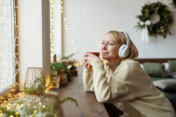 Mature woman enjoys the Christmas mood, looks out the window, listens to music and relaxes