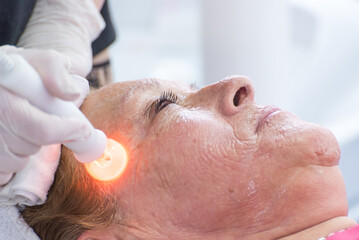 Naklejka premium Beautician performing phototherapy with LED light to adult woman in SPA salon, facial phototherapy to clean pores of the skin. Anti-aging and photo rejuvenation treatments. facial treatment.