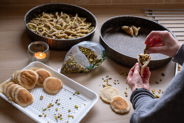 Hands holding Qatayef filled with chocolate and topped with pistachios on a wooden table, with a plate to be prepared in the oven later as Ramadan sweets.