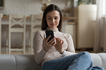 Attractive millennial female sit on couch with modern smartphone, spend leisure using new mobile...