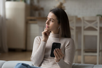 Pensive woman look into distance listen audio message via earphones on smart phone sit on sofa at...