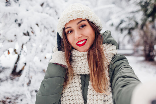 Portrait of young smiling woman taking selfie in snowy winter park wearing warm knitted hat scarf mittens. Close up