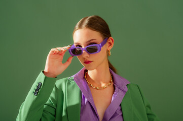 Fashionable beautiful confident woman wearing trendy purple color rectangular sunglasses, suit blazer, office shirt, chain necklace, posing on green background. Copy, empty space for text - 683555220