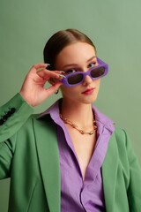 Fashionable beautiful confident woman wearing trendy purple color rectangular sunglasses, suit blazer, office shirt, chain necklace, posing on green background. Studio fashion close up portrait - 683555254