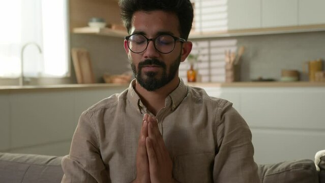 Peaceful Arabian Indian man ethnic guy closed eyes on couch at home kitchen meditate practice yoga namaste relax stress free feel calmness harmony balance comfort zen meditation spiritual awareness