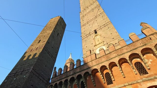Perspective bottom view of Two Towers of Bologna, symbol of city in blue sky, Italy. Asinelli tower and Garisenda tower in historic downtown with clock tower of Bologna city in Porta Ravegnana square.