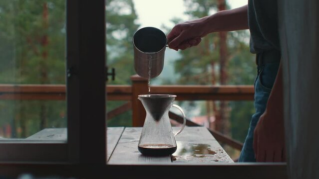 Woman pouring water into the ground coffee in pour over coffee maker with forest nature in the background.
