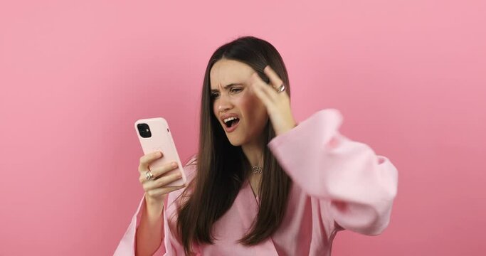 Mad and shock young brunette beautiful woman using mobile phone isolated on pink background. Yelling unhappy woman hold mobile phone. Disappointed sad upset lady horrified impressed news. WTF. Oh no