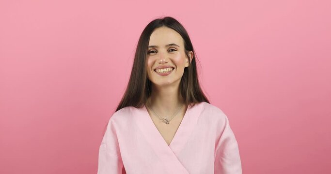 Portrait of young sexy playful brunette girl dressed in pink dress standing isolated over pink background blowing bubblegum candy bubbles and chewing gum. Woman looking at camera, laugh. Slow motion.