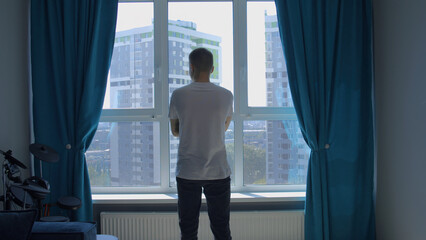 Rear view of an unrecognizable man standing at home by the window. Media. Man in white t shirt...