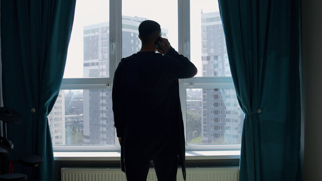 Silhouette of a business man talking on a mobile phone on the background of a window and city. Media. Successful confident businessman standing at home by the window.